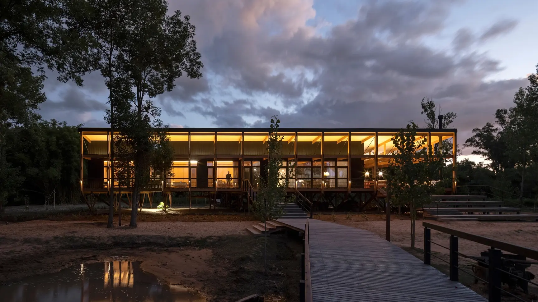 Long lit pavilion on stilts at dusk beside a boardwalk